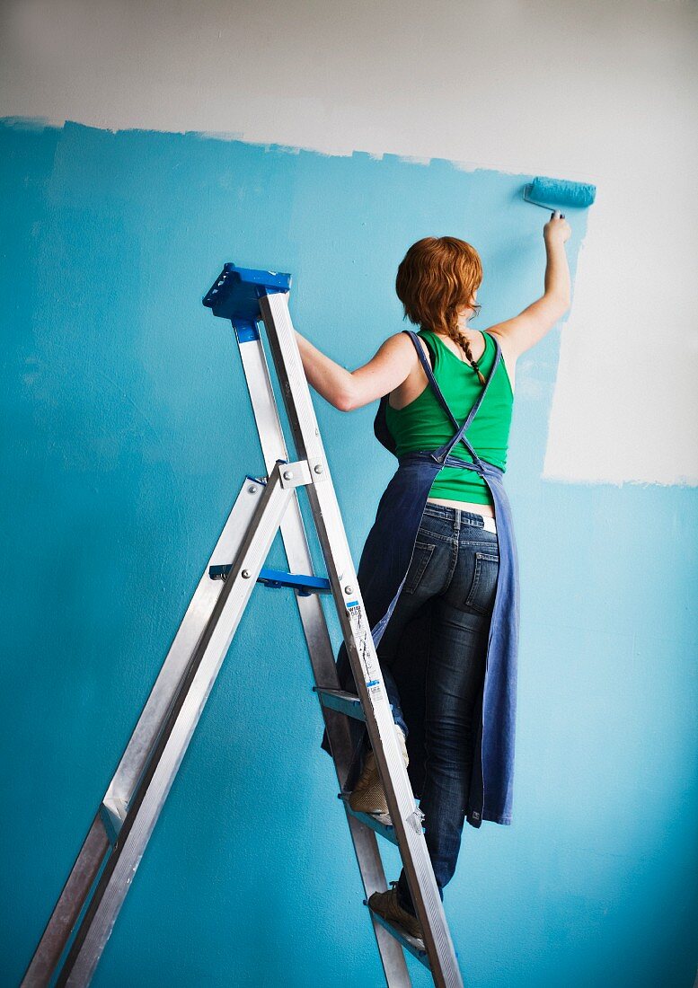 Frau auf Leiter streicht die Wand mit blauer Farbe