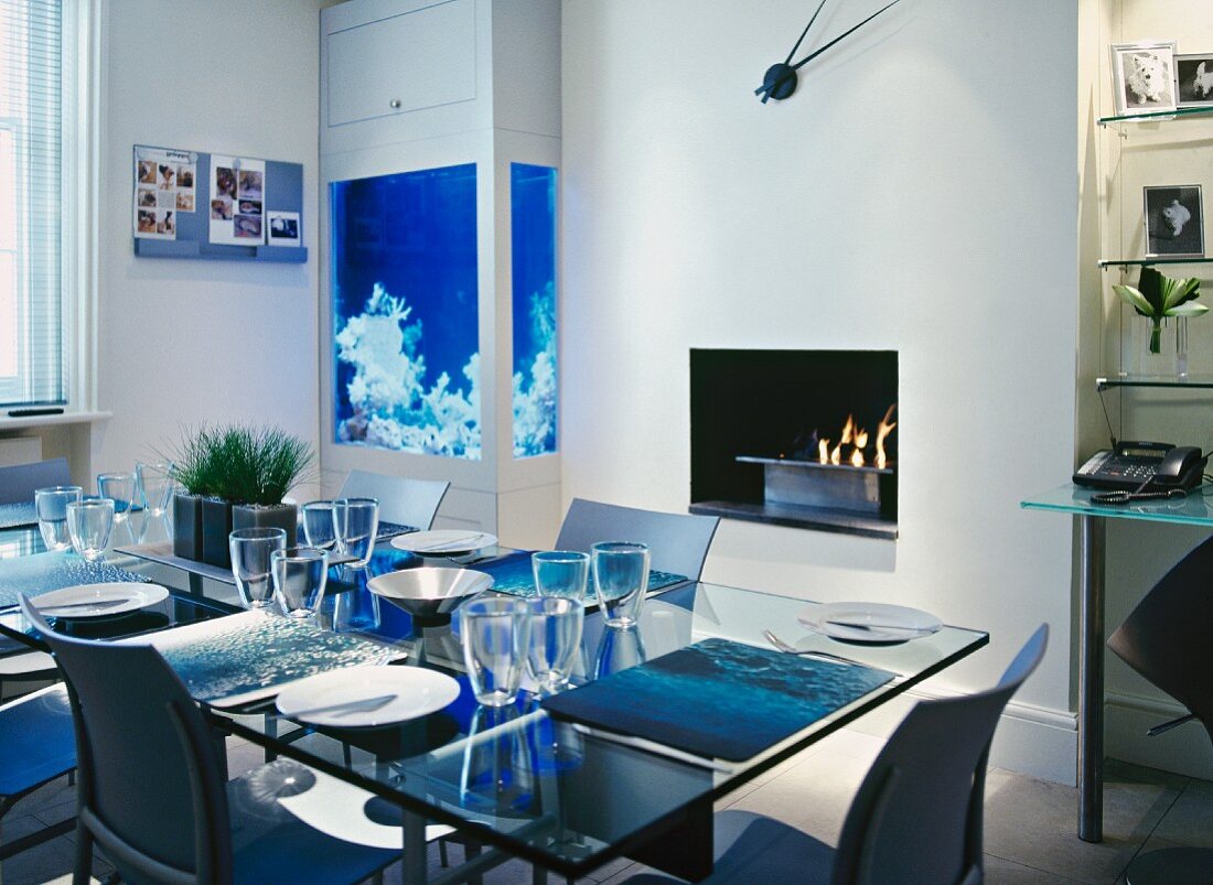 Contemporary maritime atmosphere - set glass table in front of luminous blue integrated aquarium and fire bowl in fireplace