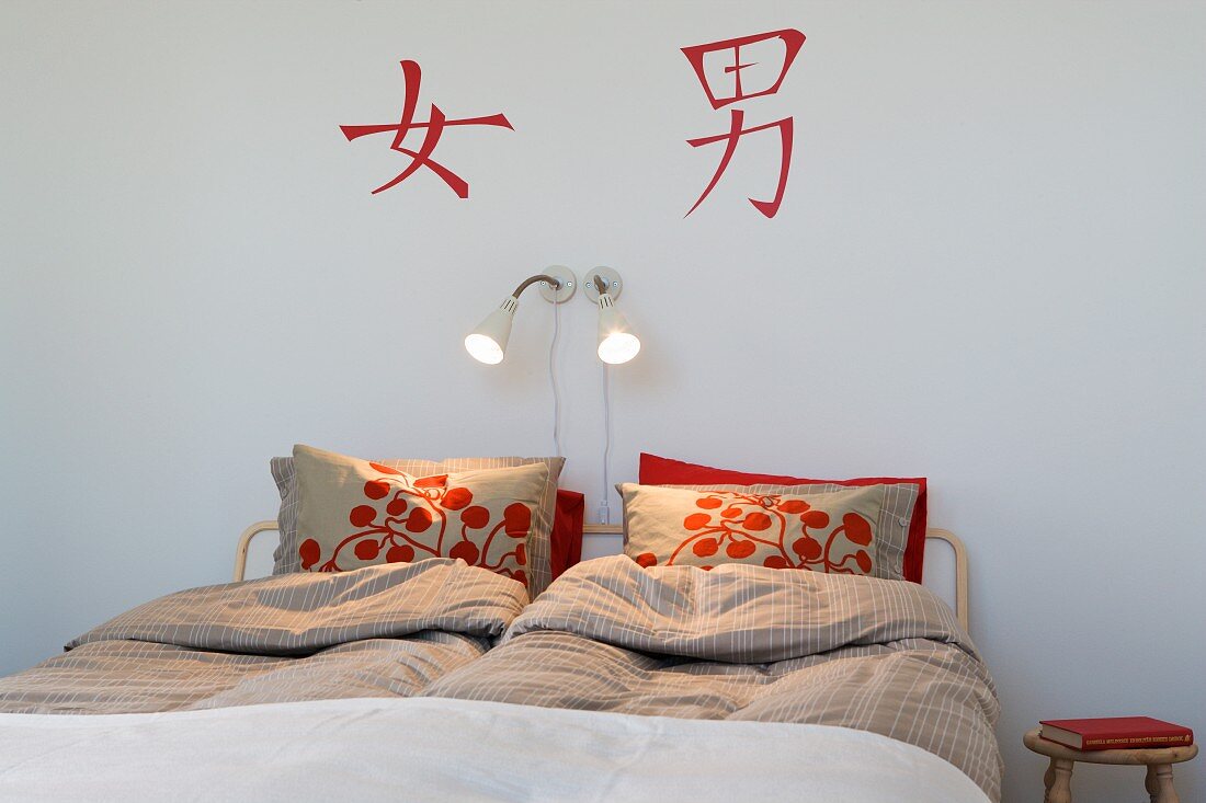 Schlichtes Schlafzimmer mit rotem Blumenmuster auf Kissen am Kopfende des Doppelbettes und rote asiatische Schriftzeichen an Wand