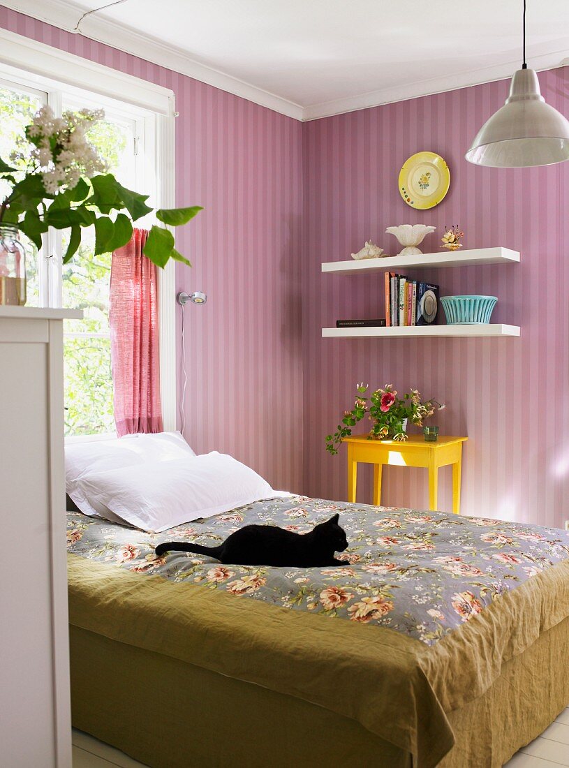 Black cat on a bed in front of a wall covered with violet, striped wallpaper