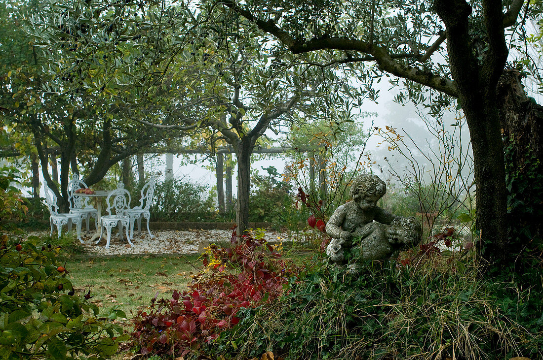 Wild garden with stone angel statue and white, vintage terrace furniture beneath pergola