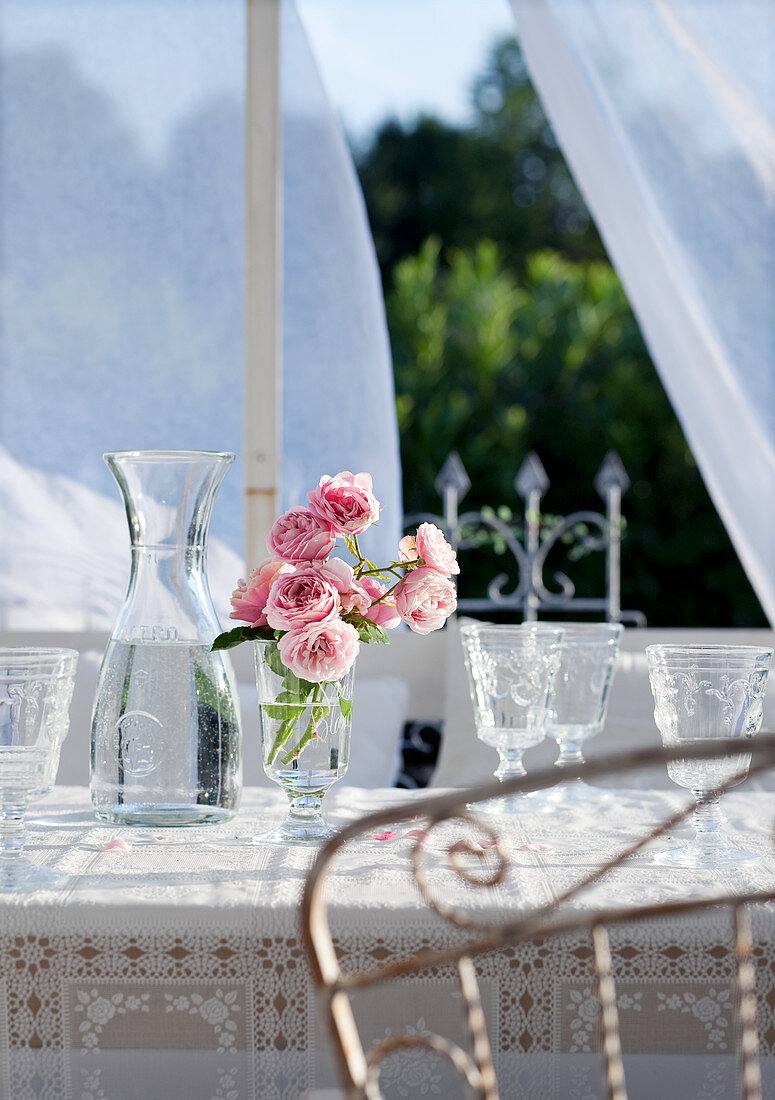 Rosen im Stielglas neben Wasserkaraffe mit Retro Weingläsern auf Terrassentisch vor transparentem Vorhang