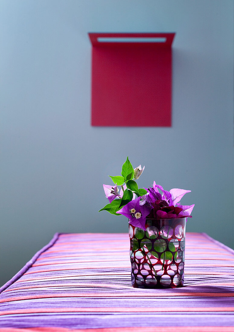 Glas mit Bougainvillea-Blüten auf gestreiftem Polster in Rot/Violett; im Hintergrund rotes Kunstobjekt vor taubenblauer Wand