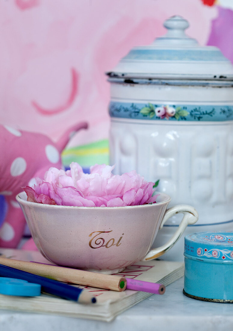 Pink flower in vintage teacup in front of vintage-style enamelled metal container