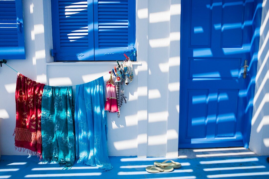 Light and shadow play on a facade of a Mediterranean home with blue window shutters and clothes hanging to dry underneath