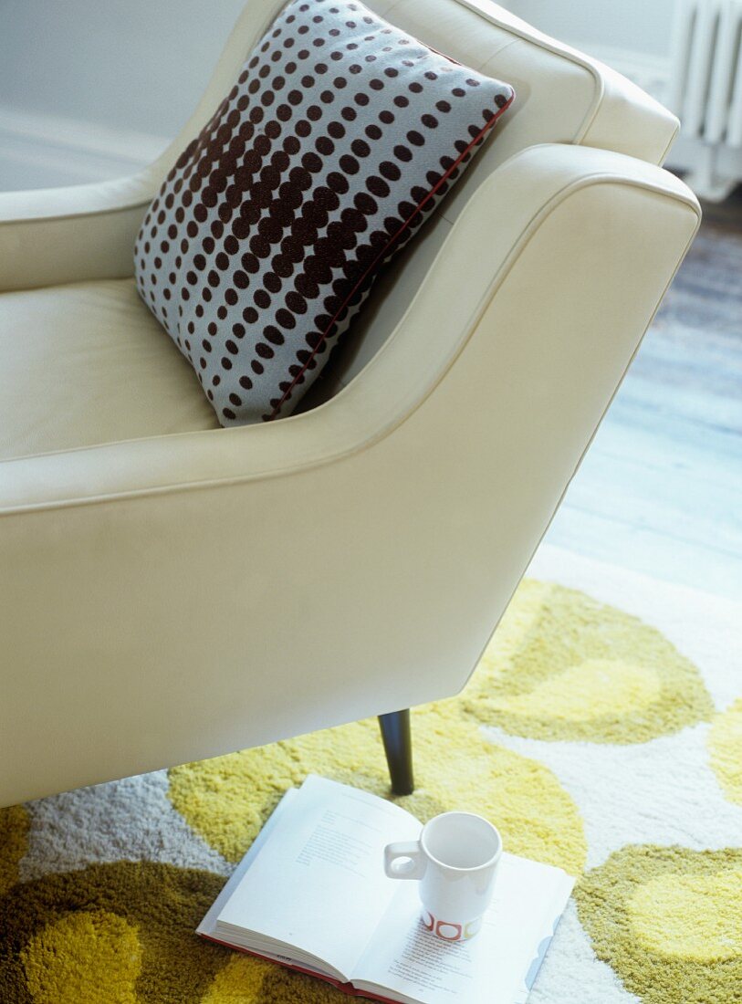 Kissen auf Sessel mit weißem Lederbezug neben aufgeschlagenem Buch und Becher auf Teppich