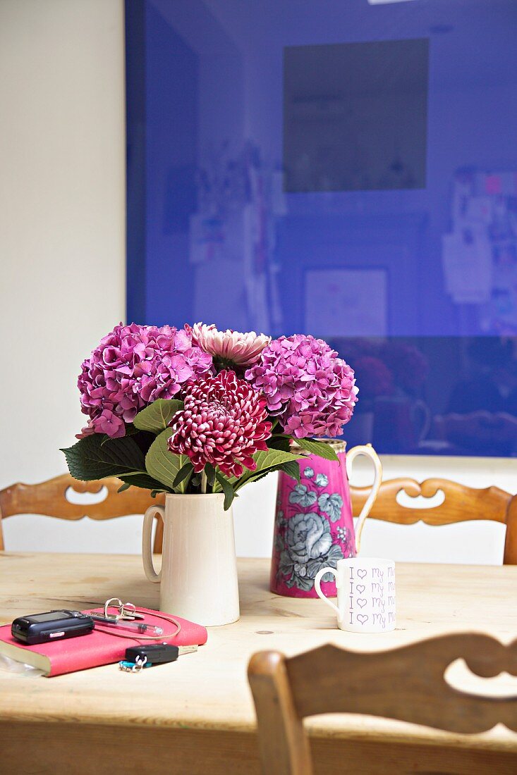 Pink violetter Blumenstrauss in weißem Keramikkrug auf Holztisch vor blauem Bild an Wand