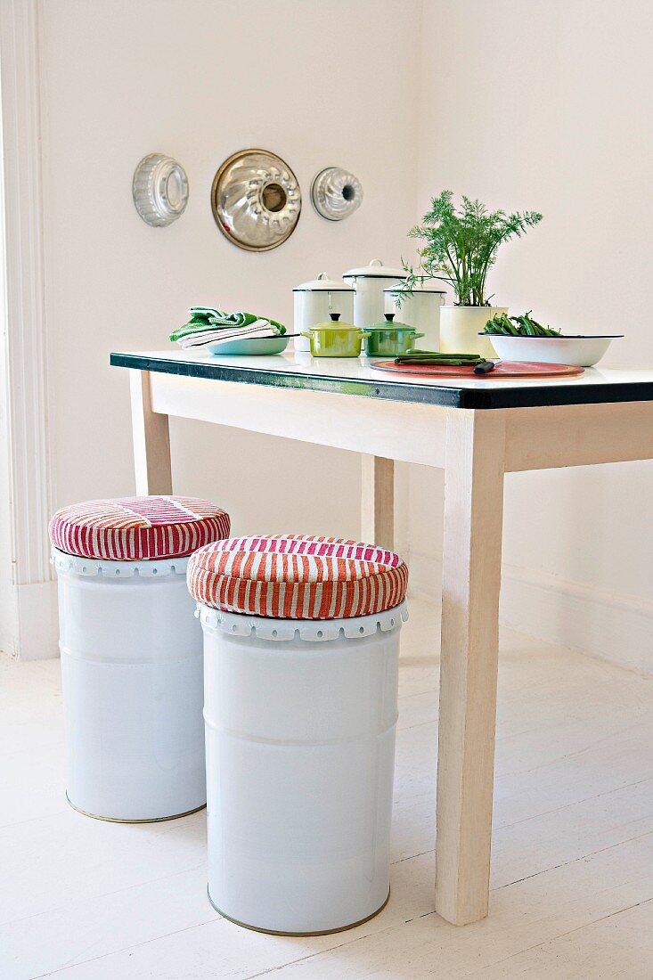 weiße Metallhocker mit Polster vor Küchentisch mit Arrangement in minimalistischer Zimmerecke mit aufgehängten Kuchenformen