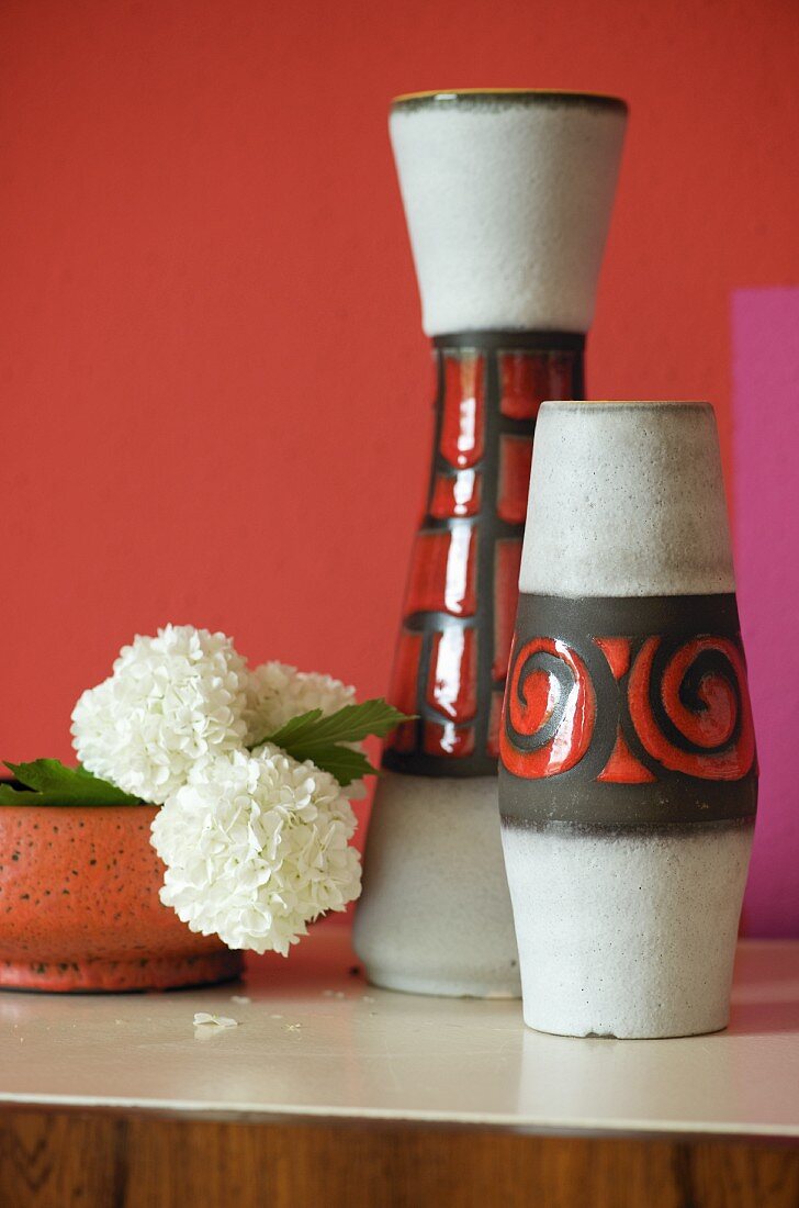 Vasenset im 70er Jahre Stil und weiße Nelken in Schale vor rot getönter Wand
