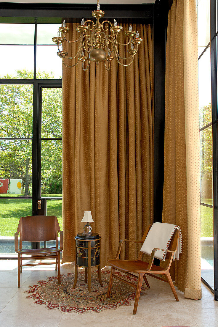 Holzstühle und Beistelltisch vor bodenlangem, hellbraunem Vorhang in Zimmerecke eines hohen Wohnzimmers mit Blick in Garten