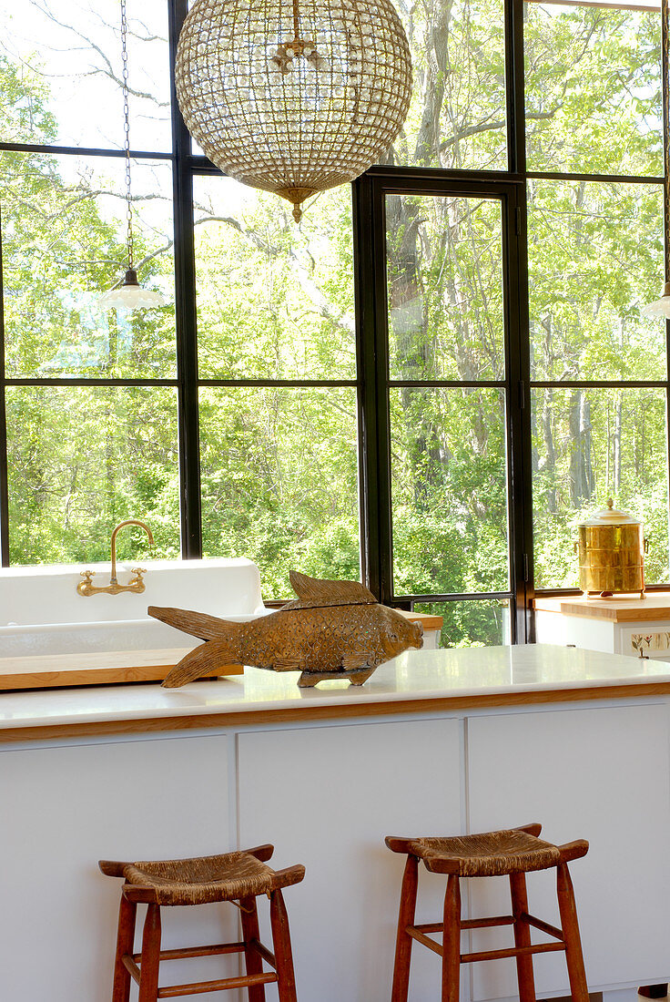 Fischobjekt aus Holz auf moderner Küchentheke und rustikale Barhocker vor Glasfenster mit Sprossen und Gartenblick