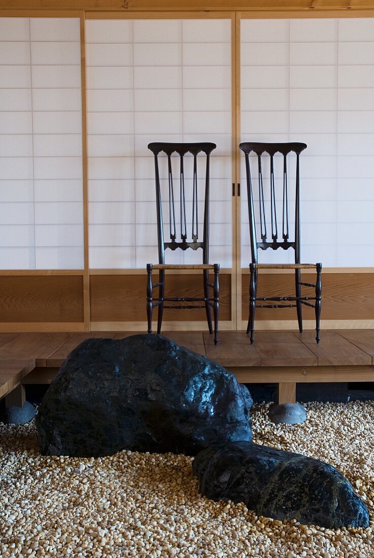 Steinfindlinge auf Kiesboden und Stuhlpaar auf Podest vor japanischer Wand