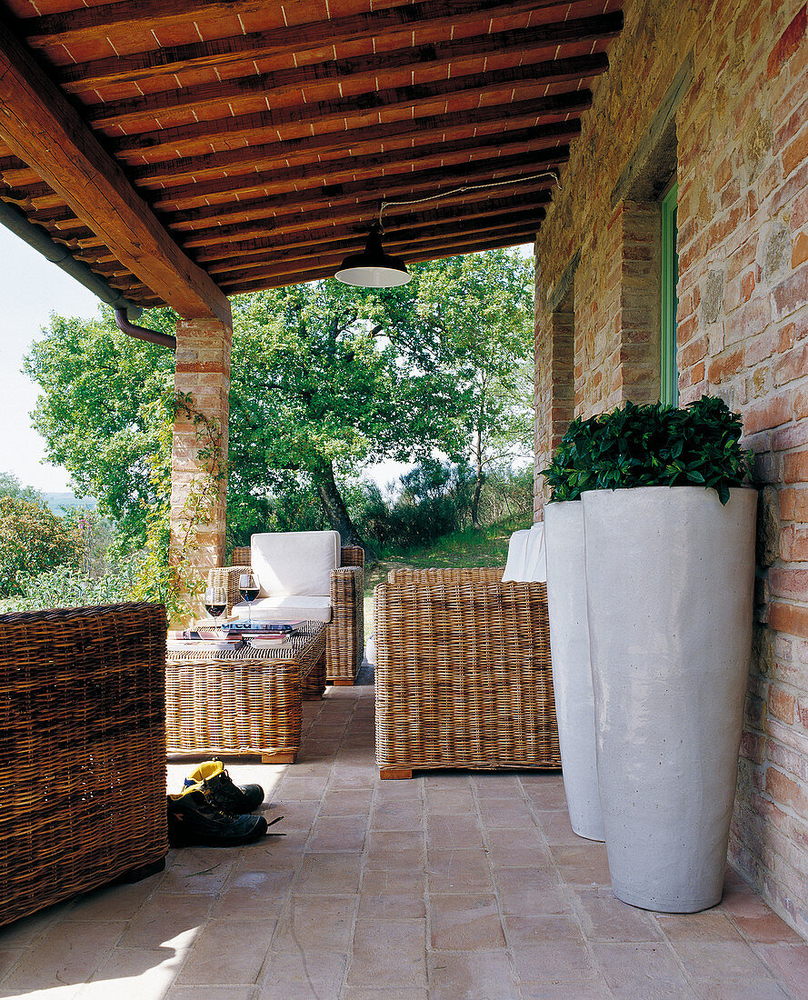 Veranda of Tuscan country house with terracotta tiles, comfortable wicker furniture and two white floor vases in foreground