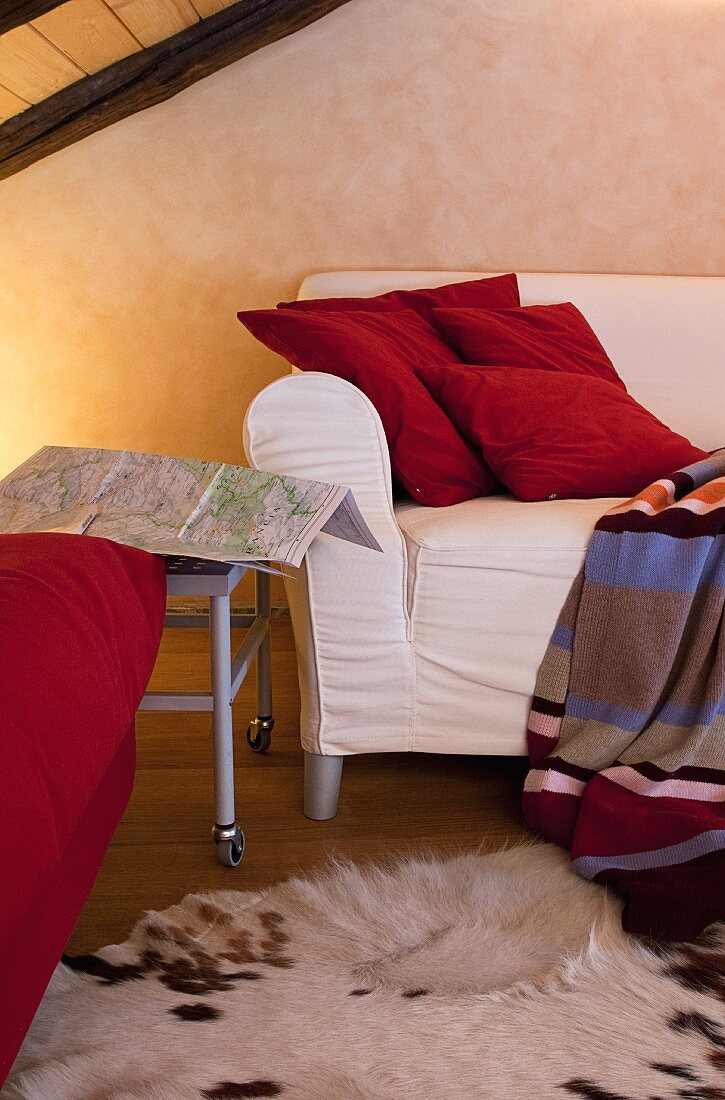Weisses Sofa unter dem Holzdach mit roten Kissen und Wolldecke im Streifenmuster; ein Tierfell als Teppich sorgt für eine gemütliche Atmosphäre