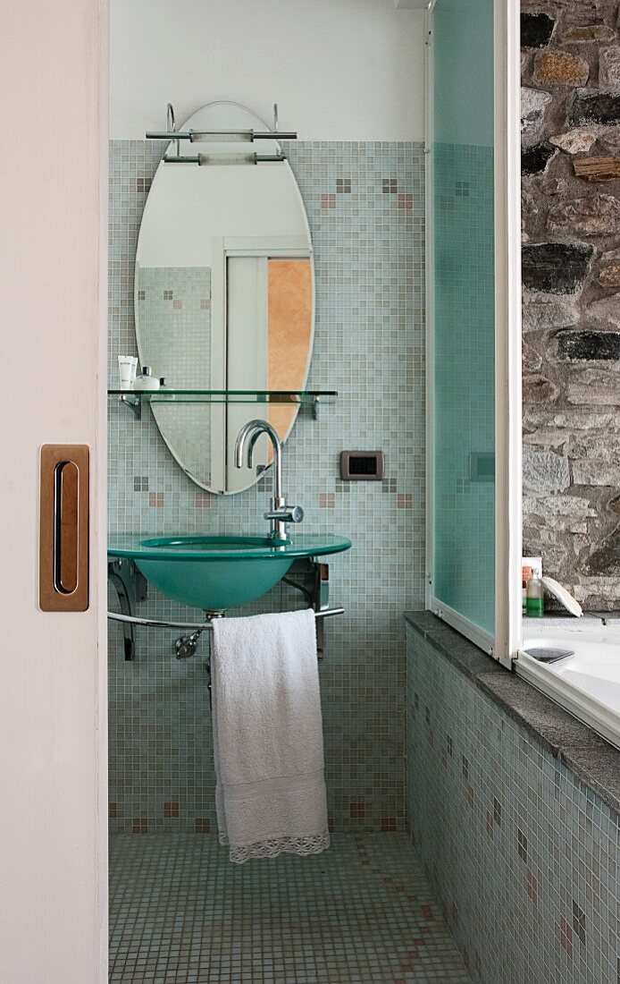 Badezimmer mit Glaswaschbecken und ovalem Spiegel; ein Schiebefenster öffnet den Blick zur aussen platzierten Badewanne