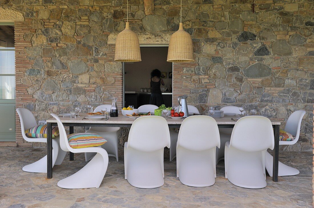 Mediterranean terrace dining area with white shell chairs and table against stone wall