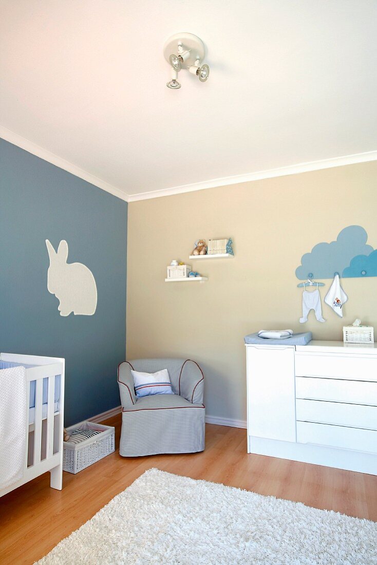 Nursery painted blue and white with rabbit motif on wall and armchair next to chest of drawers