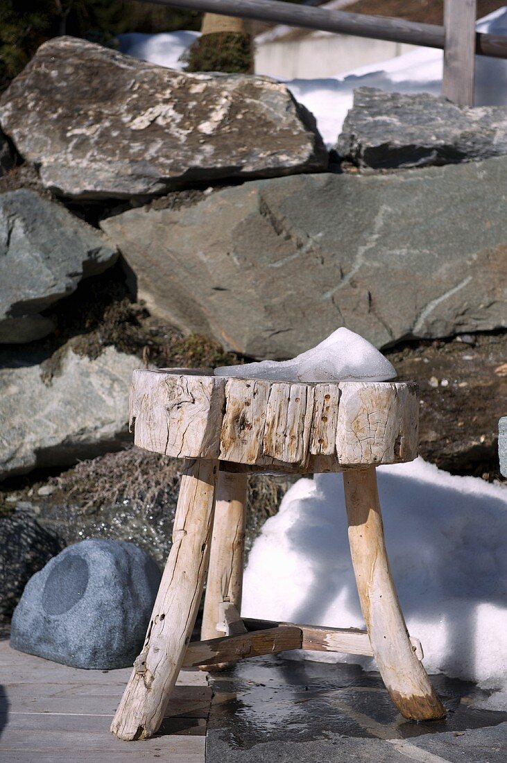 Rustikaler Holzschemel vor Felsblöcken und kleinem Schneehaufen