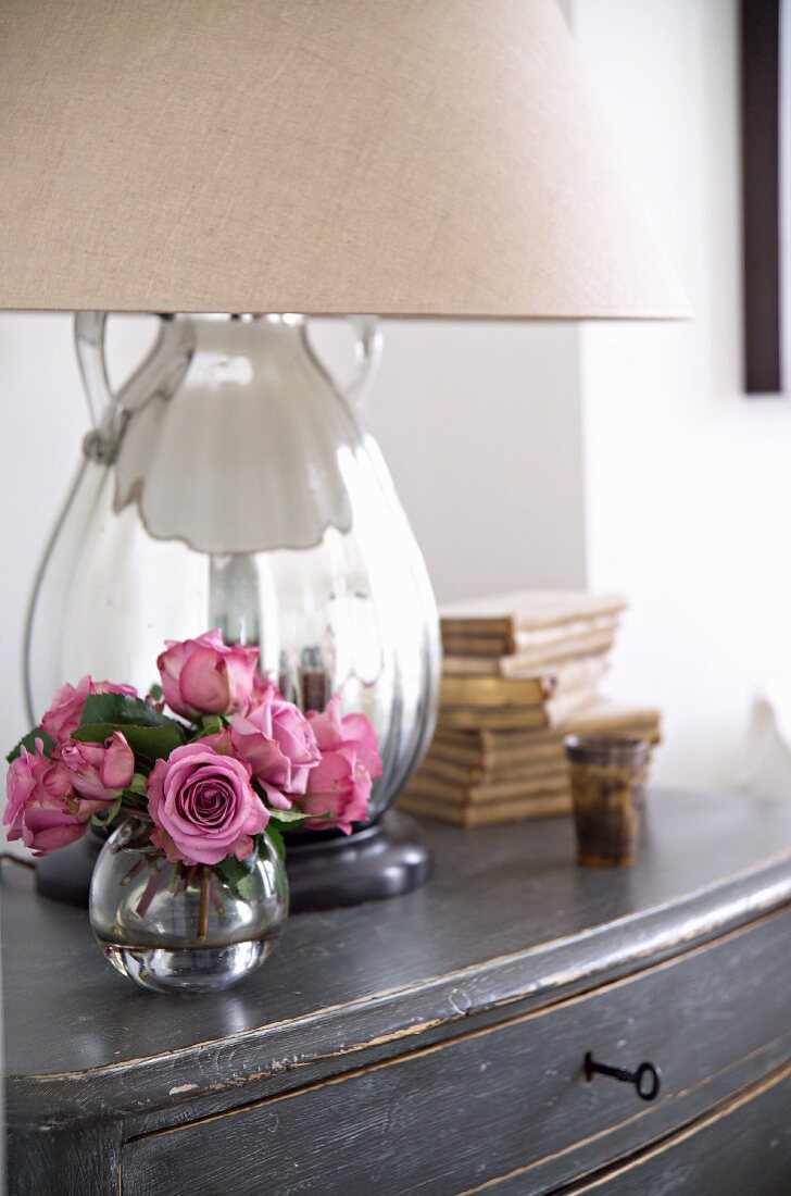 Kleiner Rosenstrauß in Glasvase vor Tischlampe mit verspiegeltem Fuß und Stoffschirm