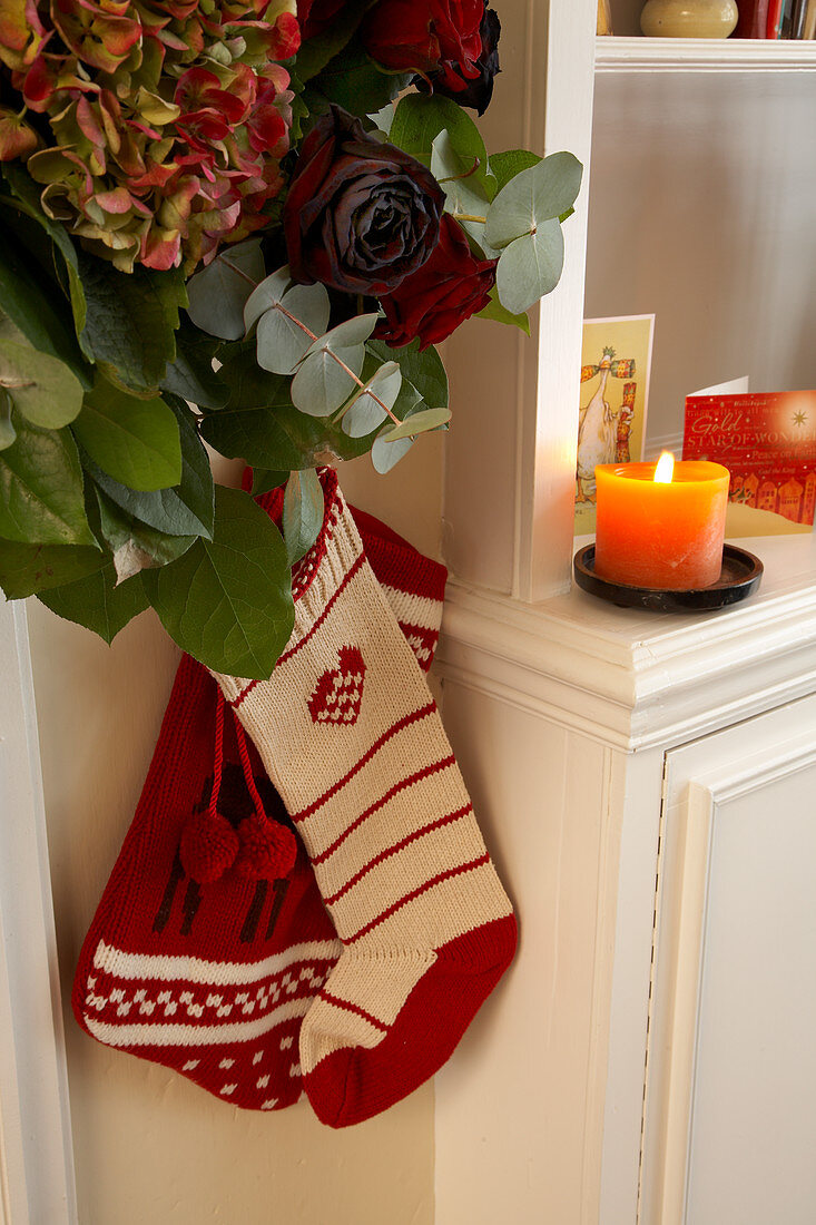 Gestrickte Nikolaussocken unter Blumendeko mit Rosen und Hortensien, daneben brennende Kerze auf Wohnzimmerschrank