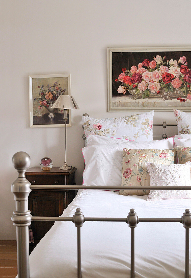 Floral pillows and scatter cushions on nostalgic metal bed below framed painting of roses