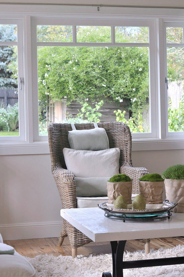 Wohnzimmer im skandinavischen Landhausstil mit Couchtisch und gemütlichem Korbstuhl vor Fensterfront mit Blick in den Garten