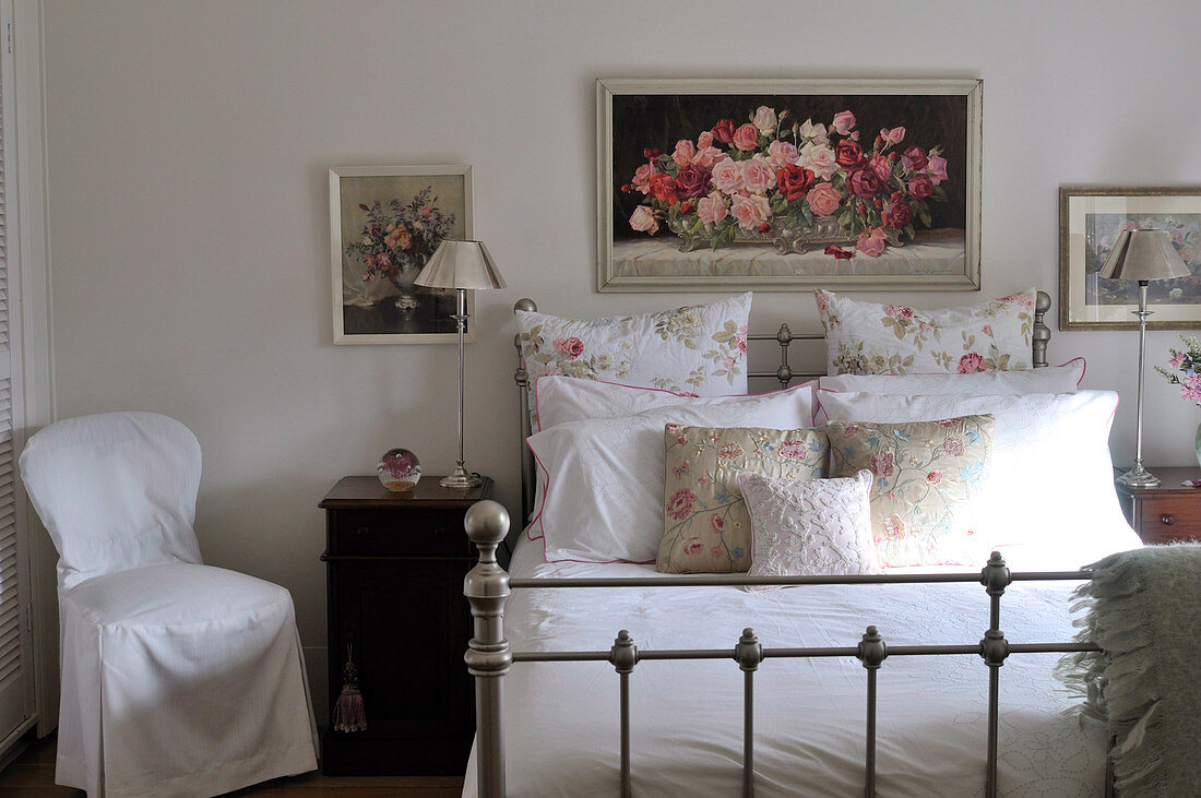 Rose-patterned cushions and pillows on metal bed with antique-inspired elements below framed painting of roses on wall