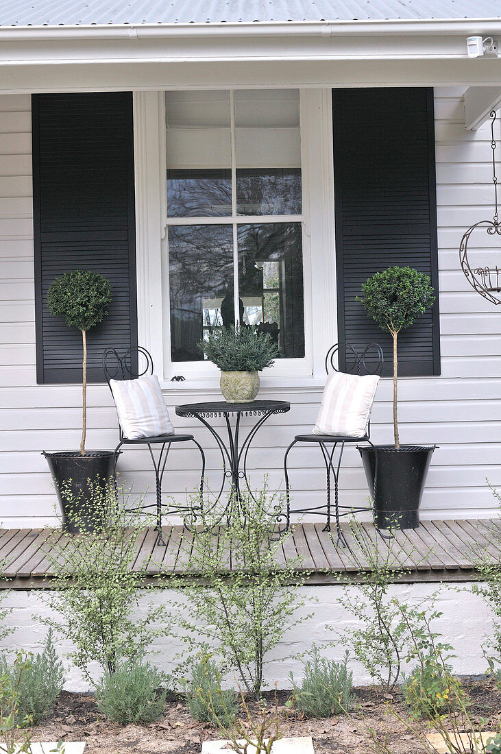 Playful bistro chairs and table in front of wooden facade and windows with dark shutters; lollipop trees in black metal planters