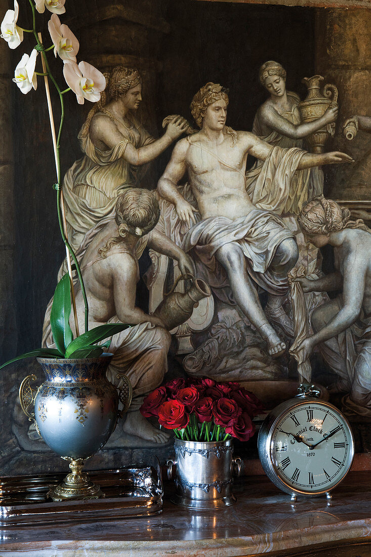 Blumen in verzierten Vasen aus Metall auf Ablage vor Bild mit antik griechischem Motiv