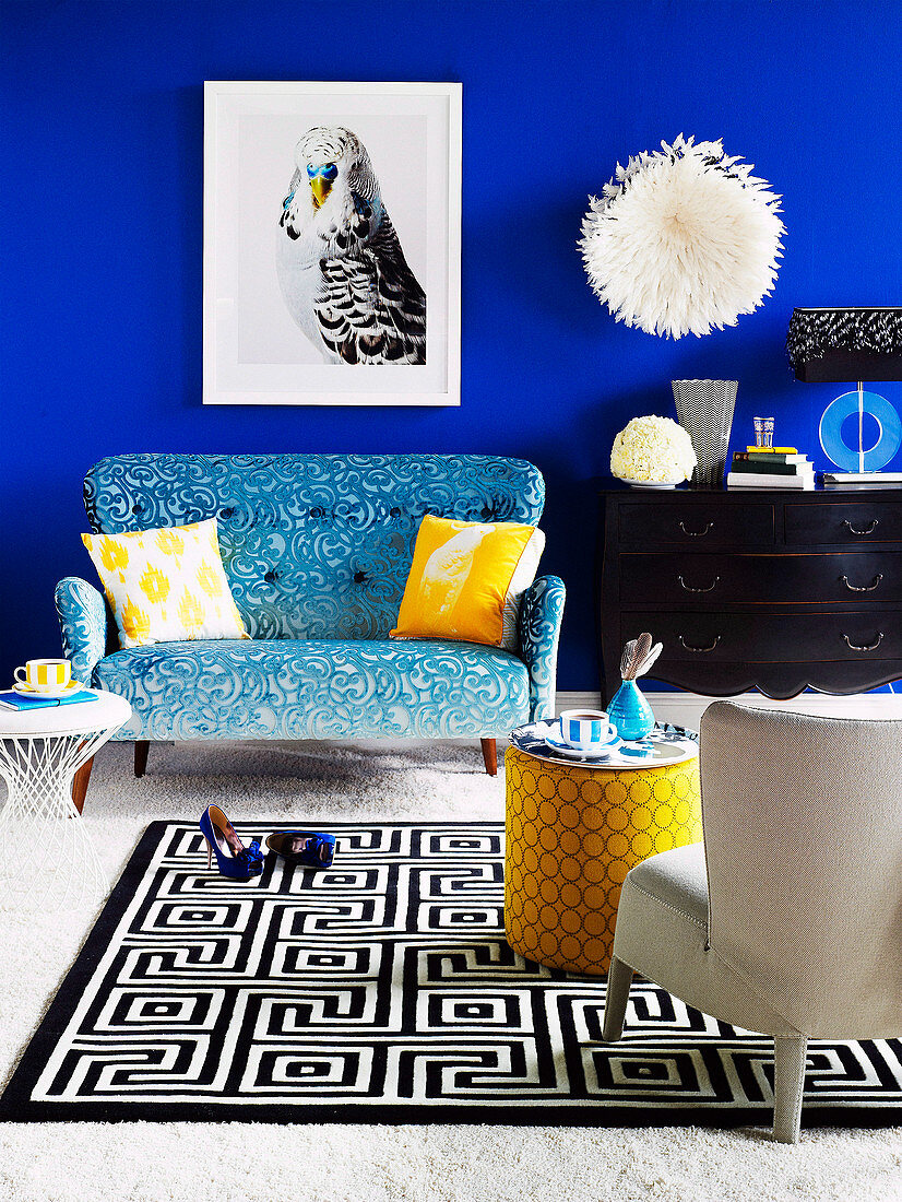 Bild mit Vogelmotiv über blauem Sofa, Kommode mit Dekoobjekten und Teppich mit schwarz-weißem Muster in einem Wohnzimmer mit blauer Wand