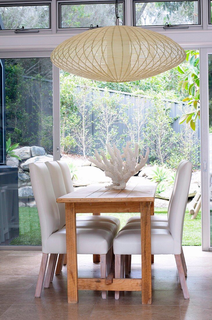 weiße gepolsterte Stühle am Holztisch und Hängelampe aus hellem Geflecht vor Terrassentür mit Blick in Garten