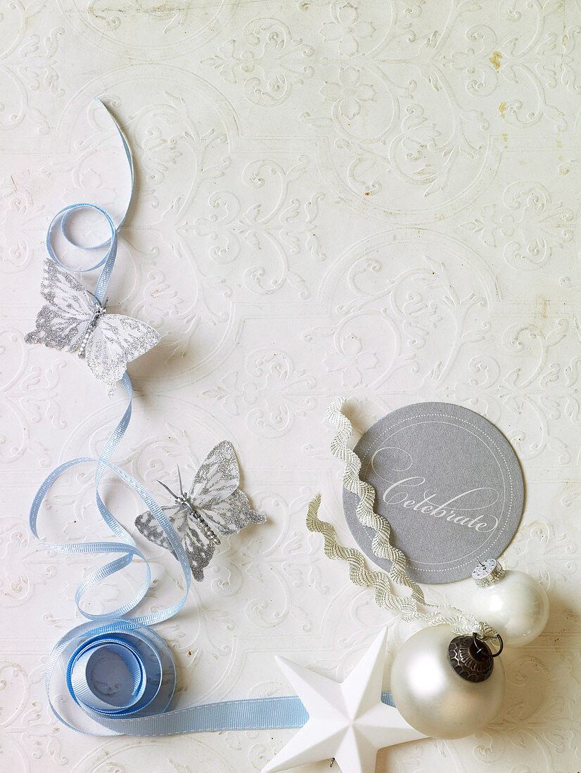 Paper butterflies next to light blue ribbon and Christmas decorations on white tablecloth with embossed pattern
