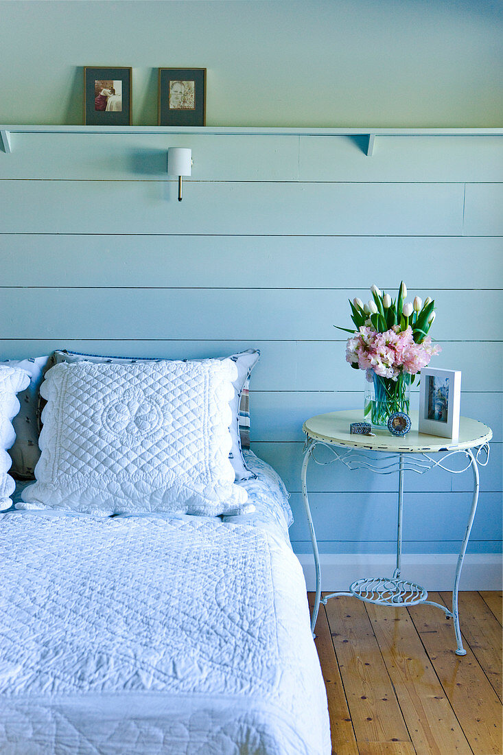 Bed with white country-style bed linen and vintage, metal side table against blue wooden wall