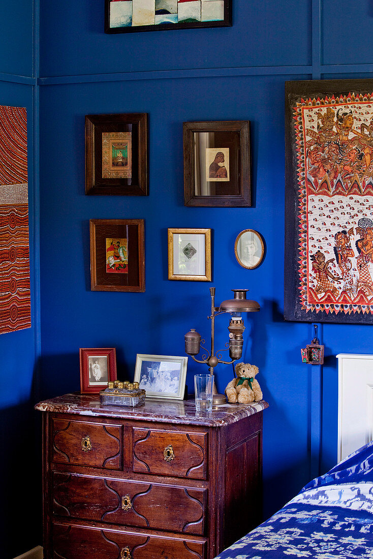 Antique chest of drawers in corner of bedroom below framed pictures on walls painted Yves Klein Blue
