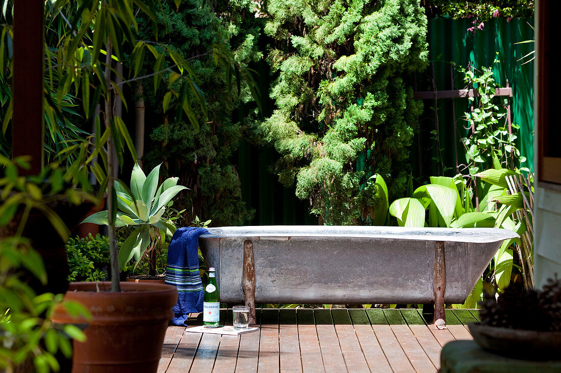 Vintage Badewanne auf Dielenboden einer sonnenbeschienenen Terrasse vor exotischem Garten