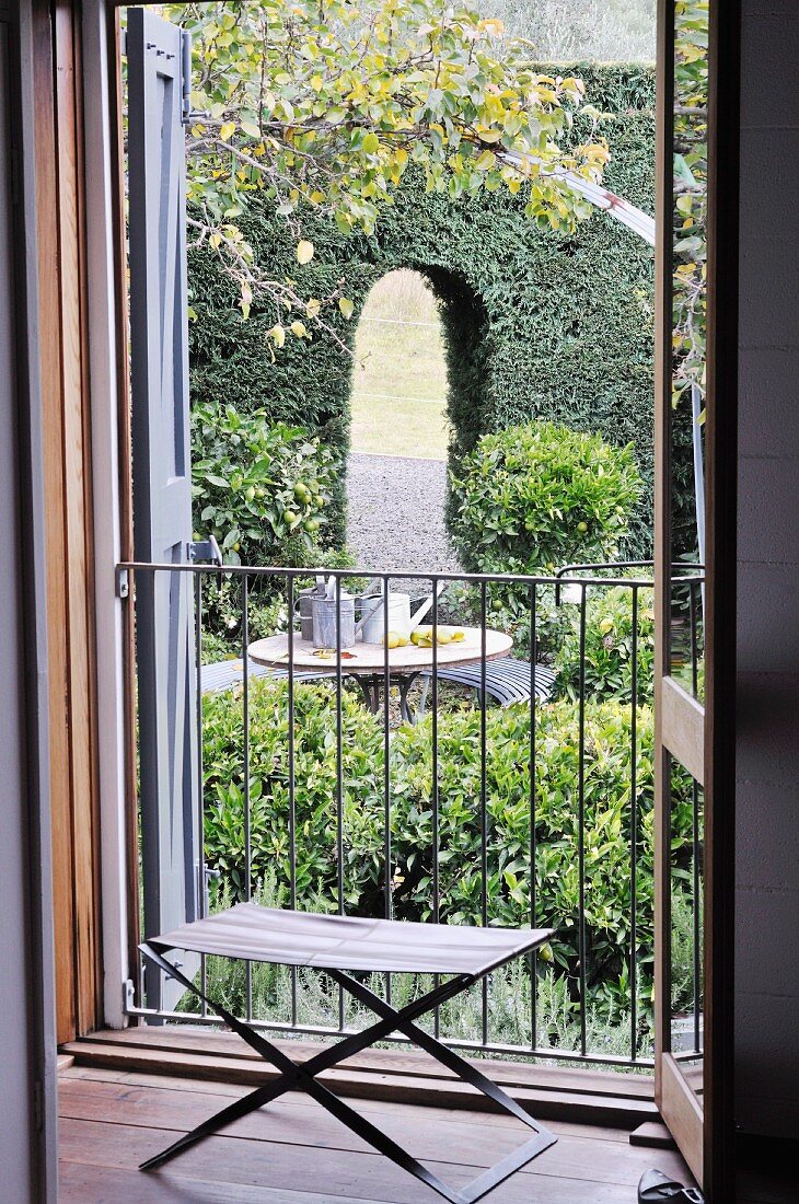 Simple folding stool in front of open French windows; view of seating area in garden in front of arched opening in tall hedge