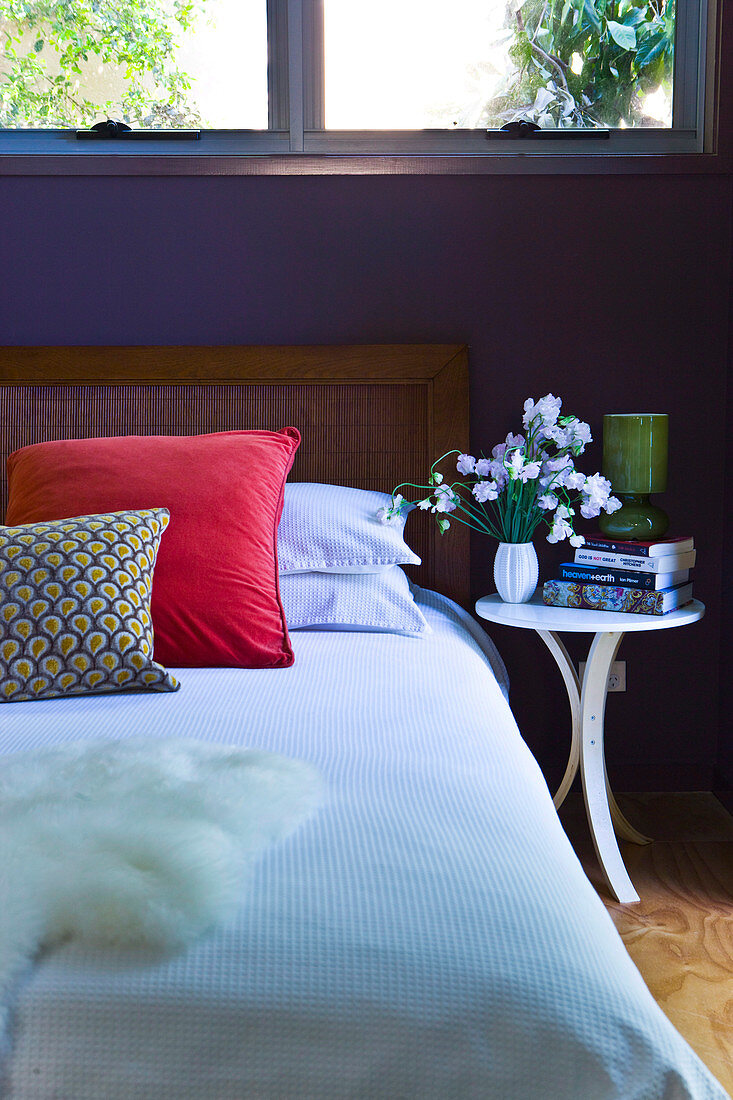 Doppelbett mit gepolstertem Kopfende unter dem Schlafzimmerfenster mit bunten Zierkissen und einem fröhlichen Blumenstrauss auf dem Nachttisch