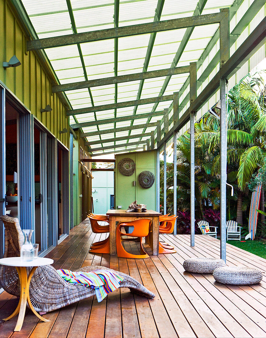 Veranda mit Glasdach, bequemer Korbliege und einem großen Holzesstisch mit orangen Kunststoffstühlen