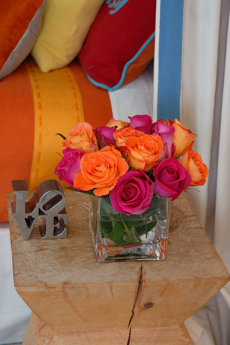 Pink und orangefarbener Rosenstrauss auf Designer-Hocker; Kissen und Bettdecke in korrespondierenden Farben im Hintergrund