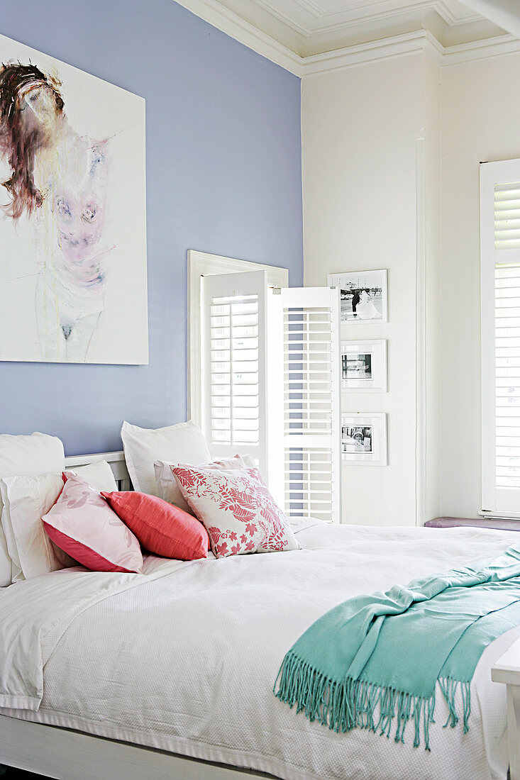 Bright bedroom with double bed against lilac wall next to window with open, white slatted shutters