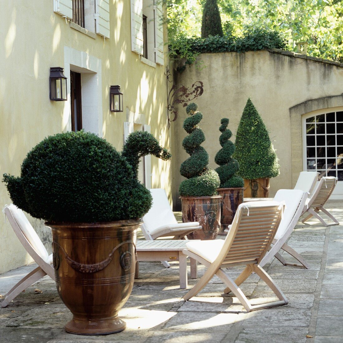 Fomgeschnittene Buchsbaumhecken in Pflanzengefässen aus Kupfer und Terrassenmöbel vor elegantem Landhaus