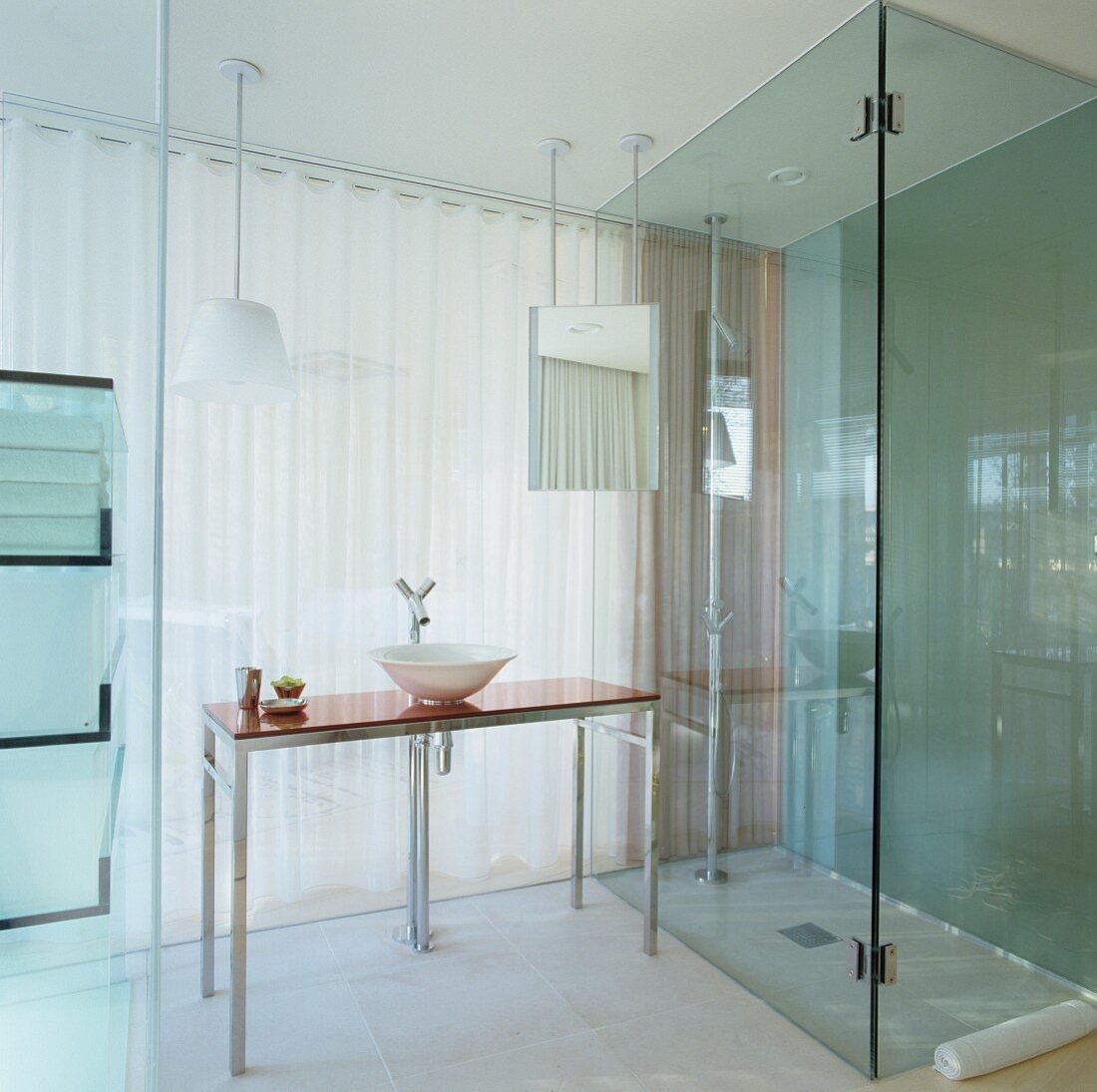 Glass ensuite hotel bathroom with floor-level shower and modern washstand with Philippe Starck tap fittings in front of translucent curtain leading to bedroom