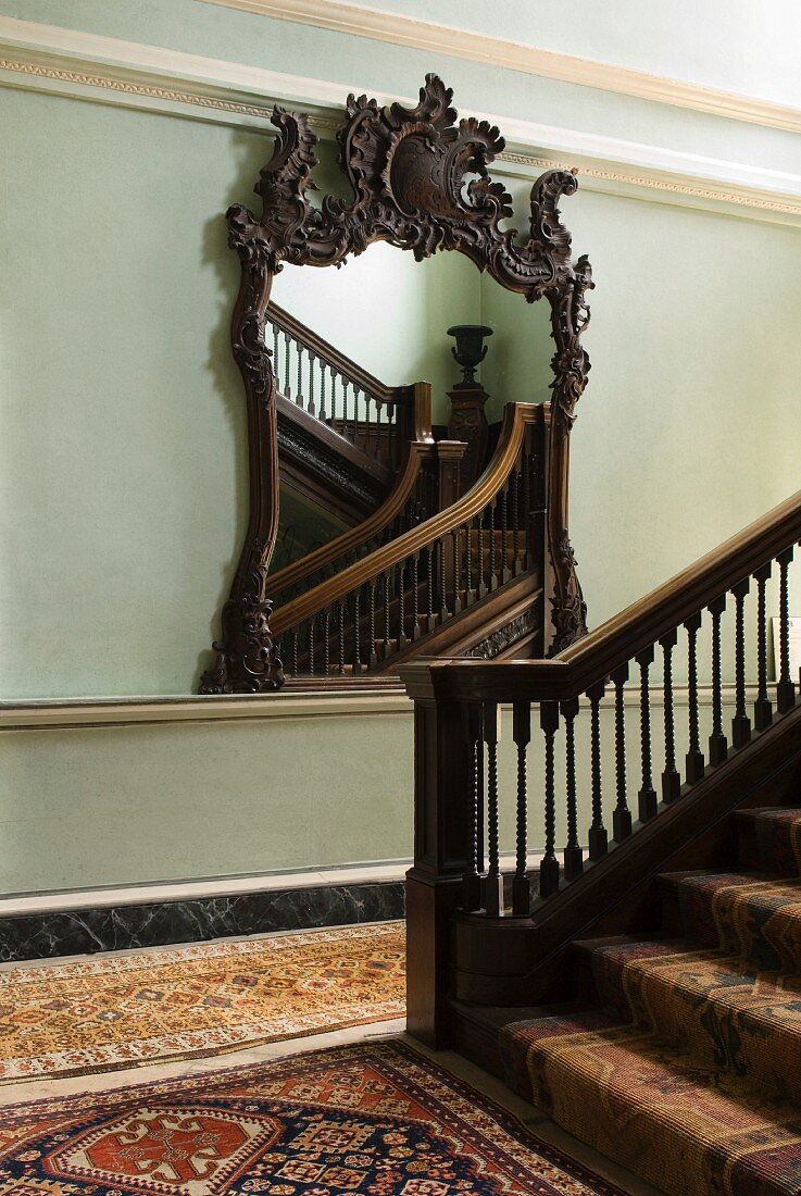 Mirror with ornately carved wooden frame, stucco elements on walls and Oriental-patterned rugs and runners in traditional stairwell