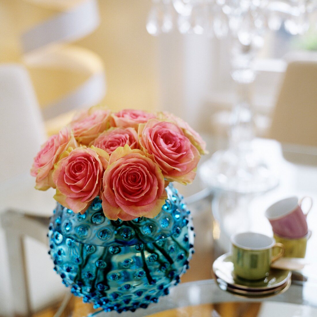 Kugelfürmige Vase aus blauem Glas mit rosa Rosen und Mokkatässchen auf Tisch