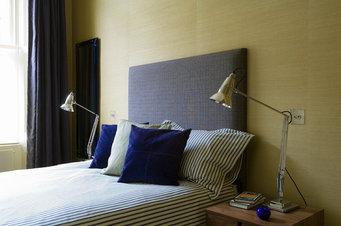 Vintage-Leuchten neben Bett mit gepolstertem Kopfteil und blauen Zierkissen vor sandfarbener Textiltapete im Schlafzimmer
