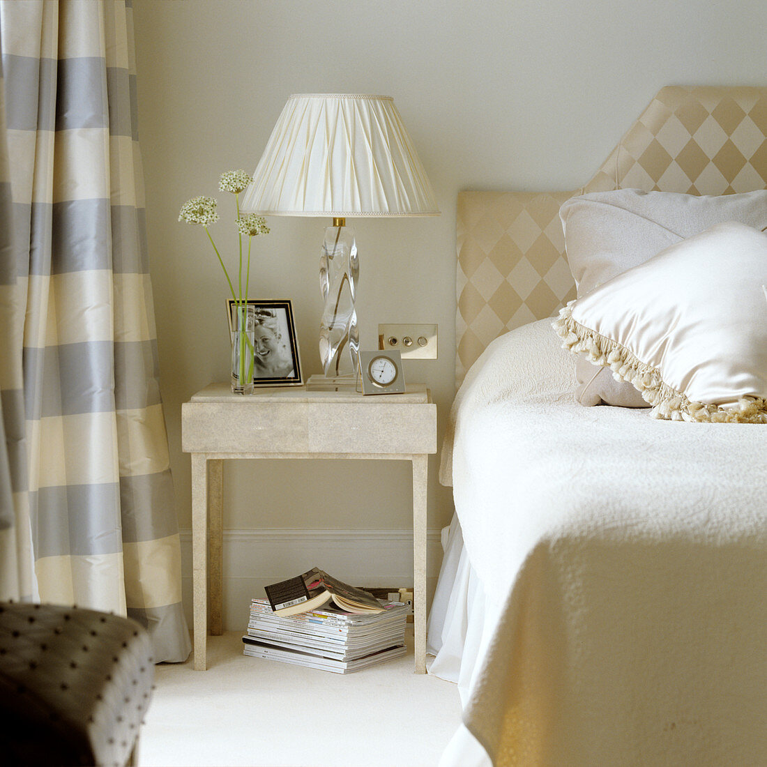 Table lamp with white fabric shade on pale side table next to bed with upholstered headboard on wall