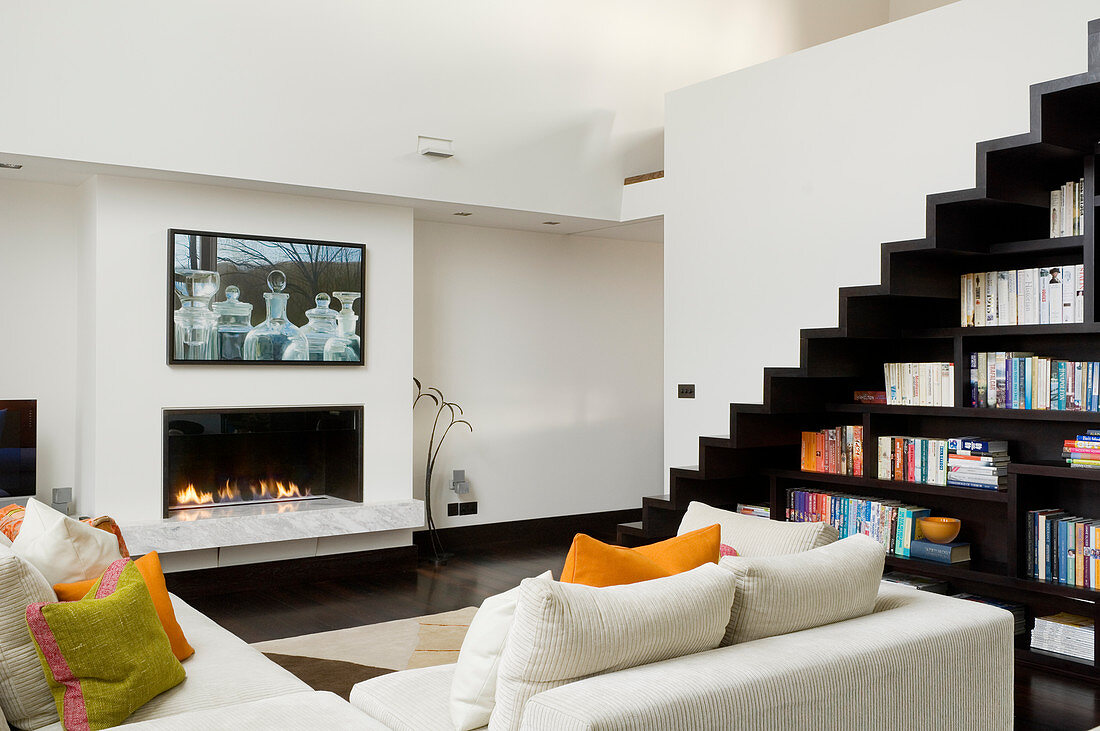 Corner sofa in front of gas fireplace in two-storey living room with zigzag staircase above custom-made bookcase