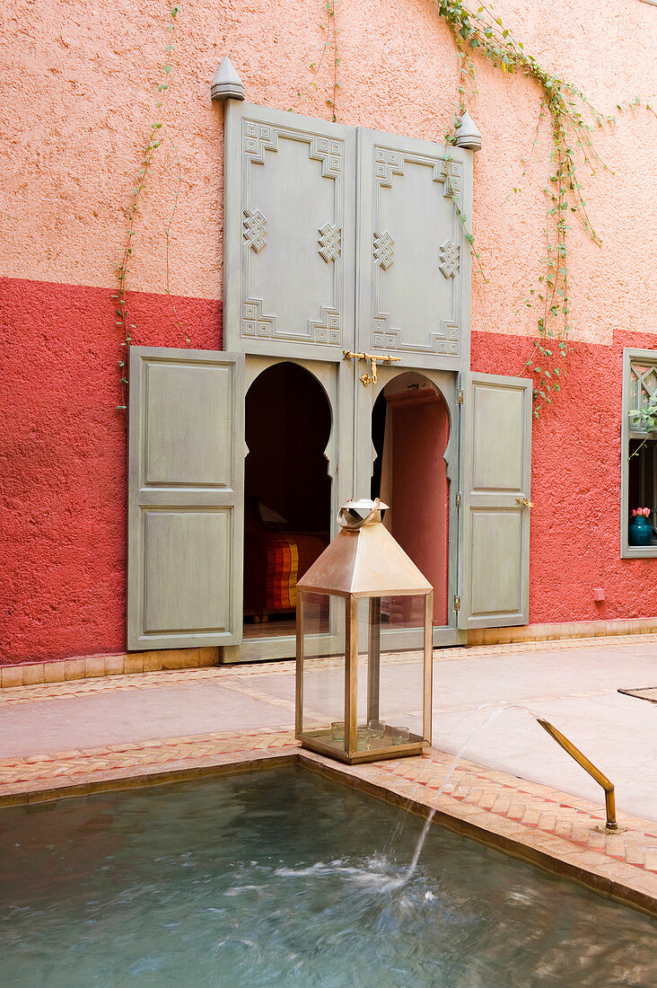 Laterne am Wasserbassin in marokkanischem Innenhof und Eingangstüren mit Spitzbogen in rotgetönter Wand
