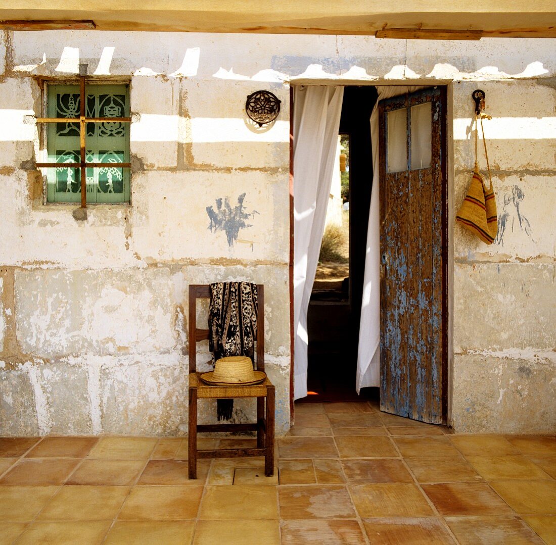 Strohhut auf rustikalem Stuhl vor unrenovierter Wand im Vorraum mit offener Tür und Blick ins Freie