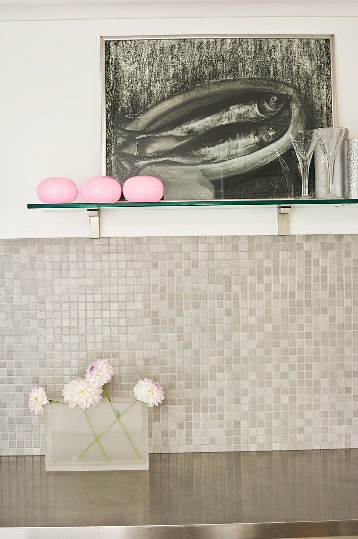 Painting and vases on wall-mounted shelf above mosaic-tiled splashback and stainless steel worksurface in modern kitchen